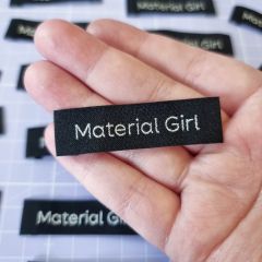 A hand holding a narrow, black sew-in label featuring the words 'Material Girl' in metallic silver