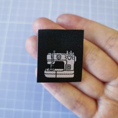 A hand holding a black woven sew-in label with the cutest little pink sewing machine on