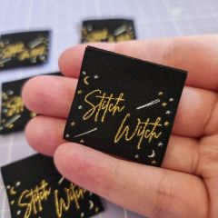 A lady's hand holding a black woven sew-in label with the words 'Stitch Witch' in metallic gold